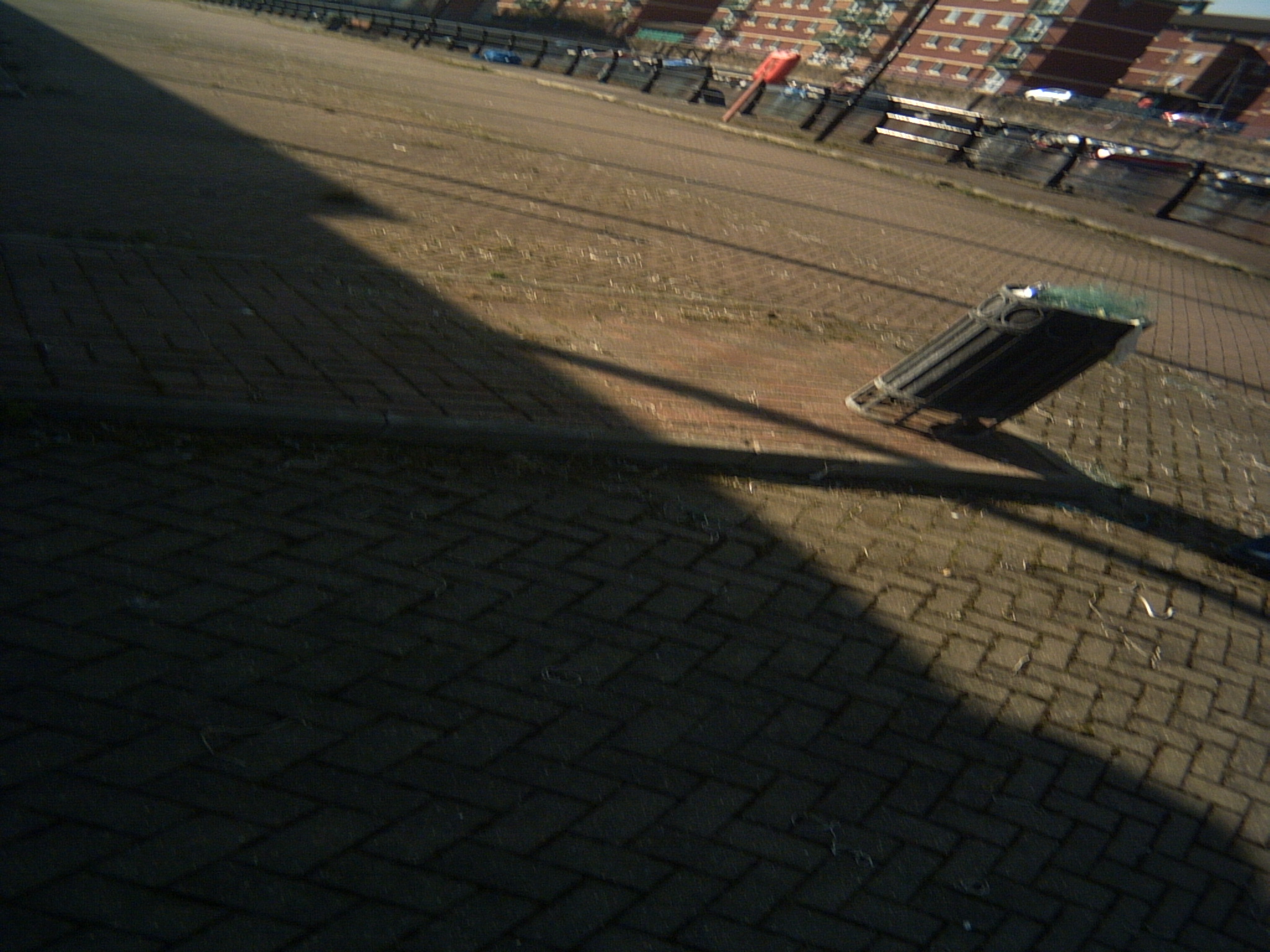 A view of Hartlepool marina, taken at a wonky angle so everything is leaning right. Most of the image is the ground, paved with maroon bricks in a herringbone pattern. The nearby pavement is almost properly horizontal, but the quay and flats in the background are leaning about 30 degrees to the right. On the pavement, towards the right of the image is a cylindrical metal bin, sheared to more than 45 degrees. The straight line of the bricks curves gently around it.